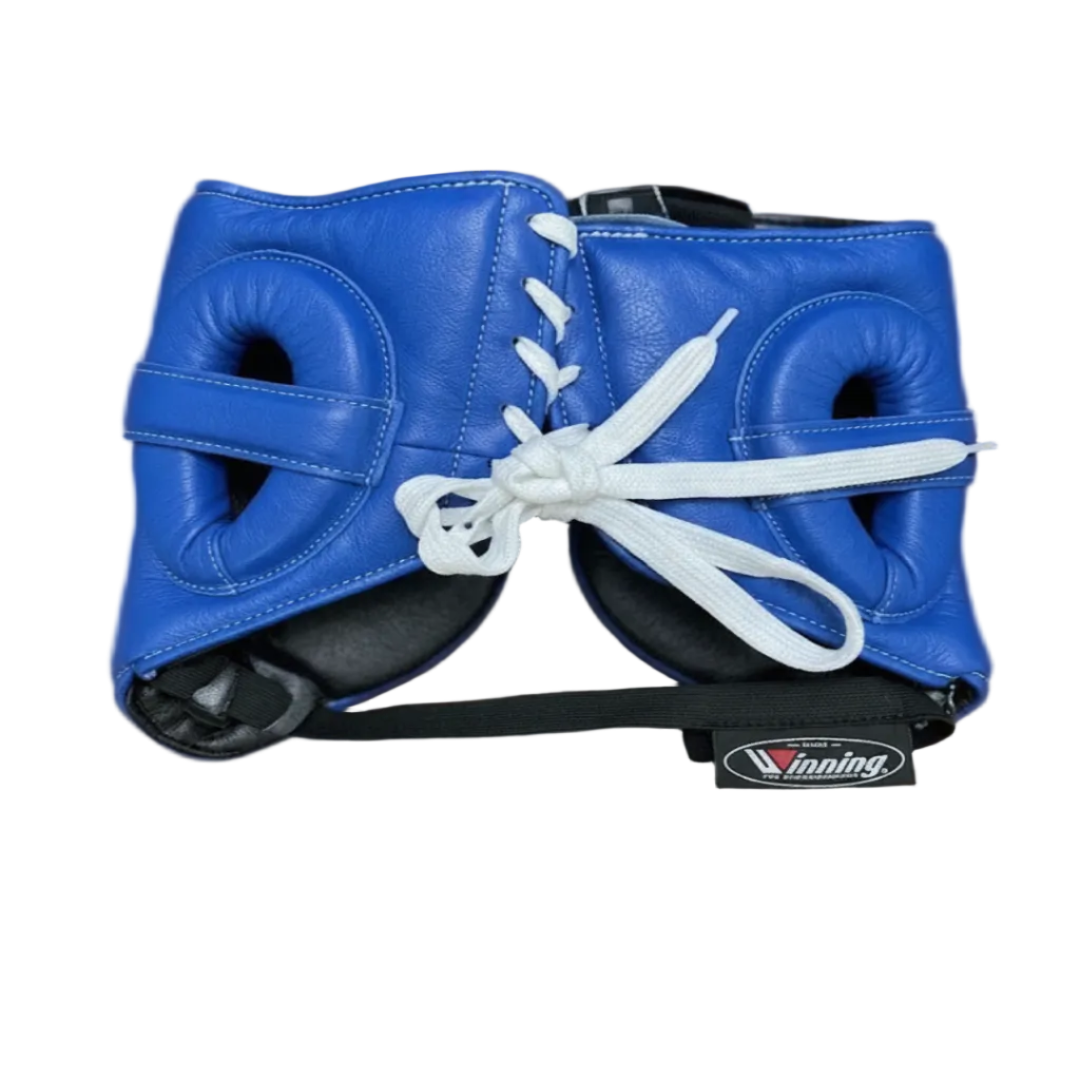 Blue boxing headgear with white laces and a black chin strap, resting on a brown couch. A small Winning brand patch is visible on the chin strap.