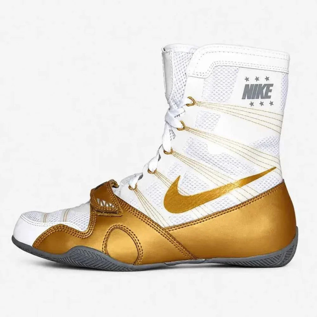 A Nike boxing boot in white and metallic gold, featuring a white mesh upper, a large gold Swoosh, and a gold toe cap, set against a light gray background.